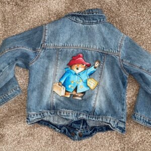 Back of denim jacket showing image of Paddington Bear, a waving brown bear wearing a blue coat and red hat holding a suitcase