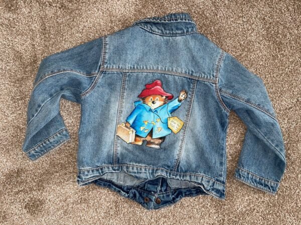 Back of denim jacket showing image of Paddington Bear, a waving brown bear wearing a blue coat and red hat holding a suitcase